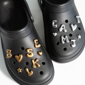 May include: A pair of black clogs adorned with decorative charms. One clog features gold charms spelling out "B S K" and other shapes. The other clog has silver charms spelling out "CAVMJ" and other shapes. The clogs are on a white background.