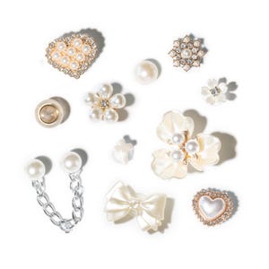 Op de afbeelding: Een assortiment decoratieve versieringen, waaronder bloemen met parelcentra, harten en een strik. De versieringen zijn in tinten wit, crème en goud, sommige met strass accenten. Een ketting met parelaccenten is ook inbegrepen.