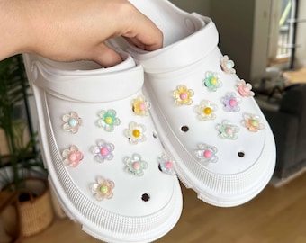 May include: White clogs decorated with colorful flower charms. The clogs have a thick sole and a strap. The flower charms are in shades of pink, yellow, green, and purple. The clogs are a casual style.