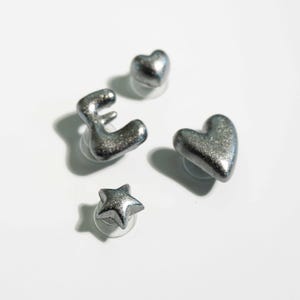 May include: Four silver-colored stud earrings in the shapes of a letter "E", a heart, a star, and another heart. The earrings have a glittery, textured surface and are set against a white background.