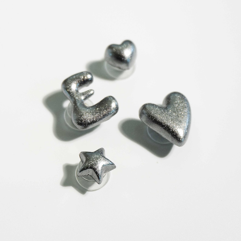 May include: Four silver-colored stud earrings in the shapes of a letter "E", a heart, a star, and another heart. The earrings have a glittery, textured surface and are set against a white background.