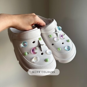 May include: White clogs adorned with star, moon, and iridescent orb charms. The clogs feature a chunky sole and are embellished with green and pearl-like accents. The text "MORE COLOURS" is visible.