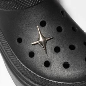 May include: Close-up of a black clog shoe with a silver star-shaped charm. The shoe has multiple round holes and a textured surface. The charm is centered on the shoe and has a shiny, metallic appearance.