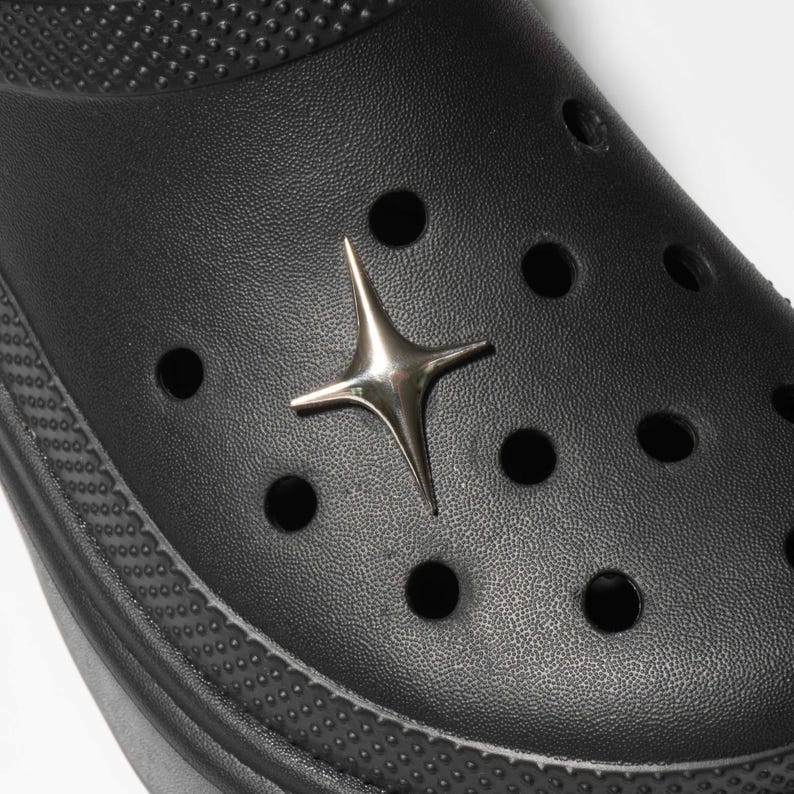 May include: Close-up of a black clog shoe with a silver star-shaped charm. The shoe has multiple round holes and a textured surface. The charm is centered on the shoe and has a shiny, metallic appearance.