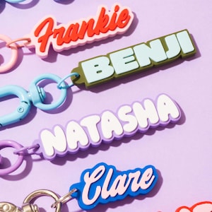 May include: A selection of name keyrings in a bubble font. The keyrings display names like "Frankie", "Benji", "Natasha", and "Clare" in bright colours including red, blue, and purple. Each keyring has a metal clasp and a chain.