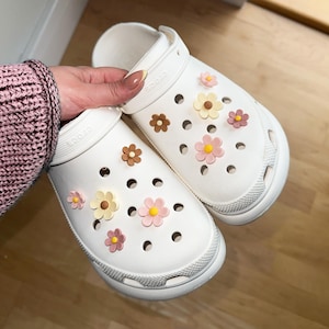 Puede incluir: Zuecos blancos decorados con abalorios de flores de colores. Las flores son rosas, amarillas y marrones. Los zuecos tienen una punta redondeada y una suela gruesa. Una persona sostiene los zuecos.