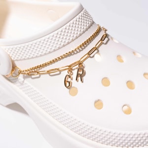 May include: A white clog shoe with a gold chain and charm. The gold chain is draped across the top of the shoe and features the letters "G" and "R" as charms. The shoe has multiple holes and a textured surface.