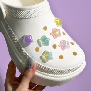 May include: White clog shoe with iridescent star-shaped charms. The charms are in various colors, including pink, purple, blue, and yellow.
