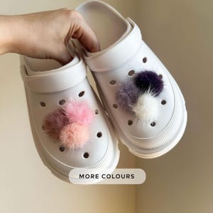 May include: White clogs adorned with fluffy pom-poms in shades of pink, peach, and purple. The clogs feature a classic design with ventilation holes and a pivoting heel strap. The image includes the text "MORE COLOURS".