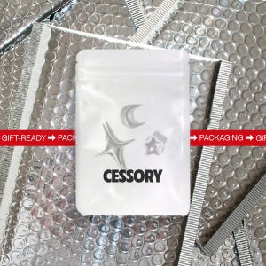 May include: A clear plastic bag containing silver-colored celestial-themed accessories: a crescent moon, a starburst, and a five-pointed star. The bag is labeled "CESSORY" in bold black letters. The background is a silver bubble wrap.