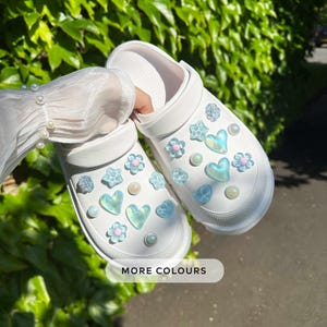 Könnte beinhalten: Weiße Clogs, verziert mit irisierenden Herz-, Stern- und Blumenanhängern sowie Perlenakzenten. Die Clogs werden vor einem Hintergrund aus grünem Laub gezeigt. Der Text "MORE COLOURS" wird unten angezeigt.