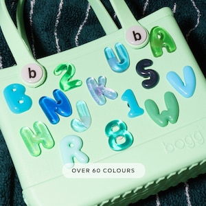 Könnte beinhalten: Hellgrüne Tragetasche mit verschiedenen bunten Buchstaben- und Zahlenanhängern. Die Anhänger sind in Blau-, Grün- und Lilatönen gehalten. Der Text "OVER 60 COLOURS" ist auf der Unterseite der Tasche zu sehen.