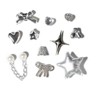 May include: A collection of silver-toned earrings in various shapes, including bows, hearts, stars, and the letter "A". Some earrings feature clear crystals or faux pearls. The earrings are displayed on a white background.