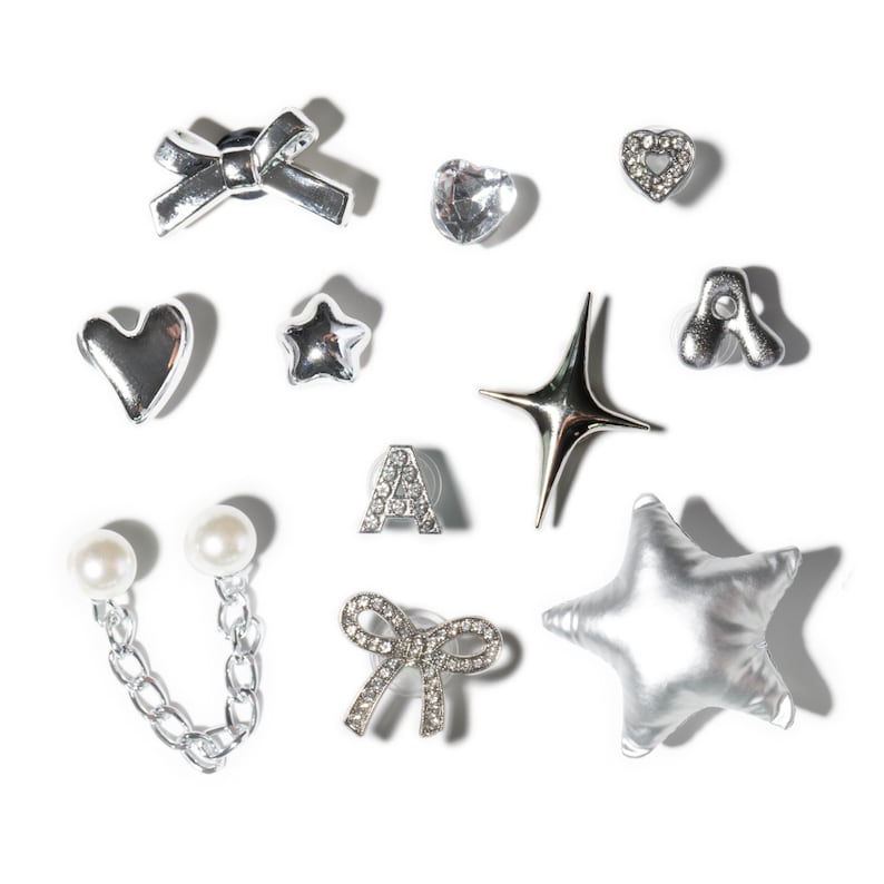 May include: A collection of silver-toned earrings in various shapes, including bows, hearts, stars, and the letter "A". Some earrings feature clear crystals or faux pearls. The earrings are displayed on a white background.