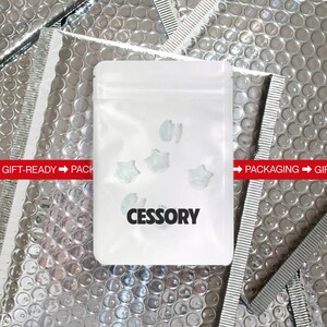 Op de afbeelding: Een witte plastic zak met het woord "CESSORY" in het zwart gedrukt. De zak bevat verschillende kleine, stervormige, lichtblauwe objecten. De achtergrond is een zilverkleurig bubbeltjesplastic materiaal.