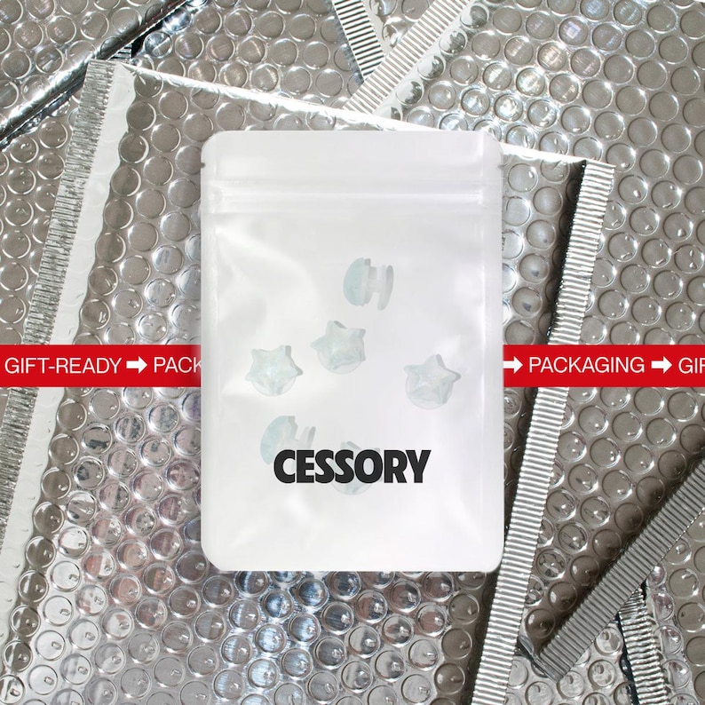 Op de afbeelding: Een witte plastic zak met het woord "CESSORY" in het zwart gedrukt. De zak bevat verschillende kleine, stervormige, lichtblauwe objecten. De achtergrond is een zilverkleurig bubbeltjesplastic materiaal.
