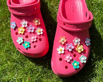 May include: Pair of bright pink Crocs clogs decorated with colorful flower charms. The clogs are made of a rubber-like material and have the word "Crocs" printed on the side. The flower charms are in various pastel colors.
