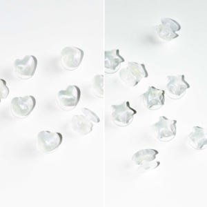 Op de afbeelding: Een assortiment iriserende hart- en stervormige kralen. De doorschijnende kralen hebben een subtiele glans, verspreid over een wit oppervlak. De hartkralen zijn afgerond, terwijl de sterrenkralen vijf punten hebben. Ideaal voor knutselen of sieraden maken.