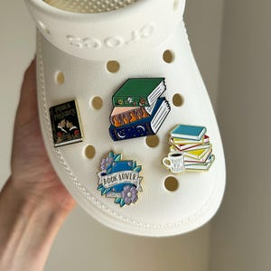 Puede incluir: Un zapato Crocs blanco adornado con pines de esmalte. Los pines presentan diseños temáticos de libros, incluyendo pilas de libros, una taza de café y un pin con el texto "BOOK LOVER". Un pin dice "Pride & Prejudice".