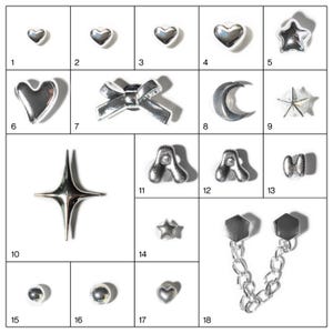 May include: A collection of silver-toned charms in various shapes, including hearts, stars, bows, moons, and letters. The charms are arranged on a white surface, showcasing their reflective qualities. Ideal for jewelry making or craft projects.