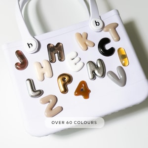 Könnte beinhalten: Eine weiße Tragetasche mit einer Vielzahl von bunten Buchstaben in Blasenform. Die Buchstaben sind in verschiedenen Farben gehalten, darunter Braun, Gold und Silber. Der Text "OVER 60 COLOURS" ist unten zu sehen.