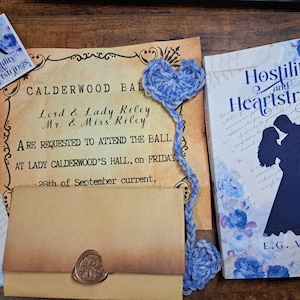 May include: A blue and white crocheted heart bookmark with a blue and white crocheted tassel. The bookmark is on top of a vintage-looking invitation for a ball. The invitation reads "Calderwood Ball, Lord & Lady Riley, Mr. & Miss Riley, Are requested to attend the ball at Lady Calderwood's Hall, on Friday 28th of September current."
