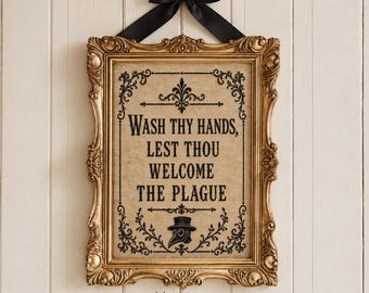 Può includere: Una targa da parete rettangolare in una cornice dorata ornata con un fiocco di nastro nero in alto. La targa presenta uno sfondo in juta color marrone chiaro con testo nero: WASH THY HANDS, LEST THOU WELCOME THE PLAGUE, e un'icona di medico.