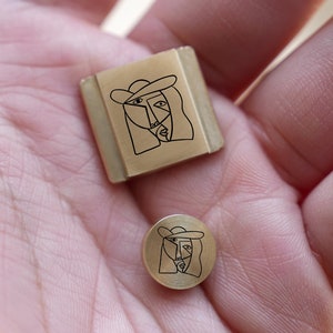May include: Two brass-coloured metal stamps, one square and one round, each featuring a minimalist line drawing of a person wearing a hat. The stamps are held in a hand, showcasing their size and design.