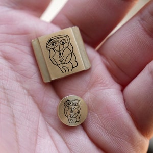 May include: Two gold-toned metal stamp designs featuring a black line drawing of a face. The larger stamp is rectangular and the smaller stamp is round.