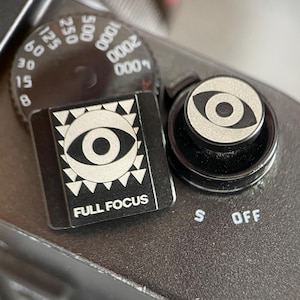 May include: A black camera hot shoe cover and a matching shutter release button, both featuring a silver eye graphic. The square hot shoe cover displays the text FULL FOCUS below the eye, while the circular shutter button has a centered eye.