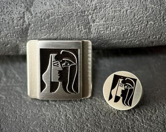May include: A silver metal cufflink with a black and white engraved image of two faces. The cufflink is square shaped and has a smooth surface.