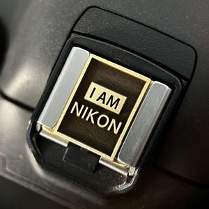 May include: Close-up of a black camera hot shoe with a gold and bronze metal plate. The plate has the text "I AM NIKON" in white lettering. The hot shoe has silver metal accents.