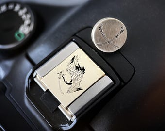 May include: A close-up of a camera accessory with a crane design and a round metal piece with crossed swords. The crane is black and white, and the metal pieces have a brushed finish. The items are on a black surface.