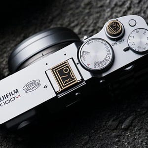 Puede incluir: Una cámara plateada Fujifilm X100VI con detalles en negro. La cámara tiene diales para la configuración y un sistema de lentes. La parte superior de la cámara tiene un interruptor de encendido/apagado y un dial con números. La cámara tiene el texto "FUJIFILM X100VI".