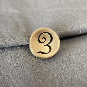 Pode incluir: Um bot&atilde;o circular de tom dourado mate com o n&uacute;mero tr&ecirc;s gravado em escuro numa fonte estilizada e curva. O bot&atilde;o tem uma superf&iacute;cie lisa ligeiramente c&ocirc;ncava e um rebordo definido, apresentando um design simples e funcional.