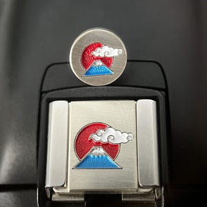 May include: A silver-coloured pin and rectangular clasp with a Japanese-inspired design. The design features a red sun, a blue mountain, and a white cloud. The pin is round, while the clasp is rectangular.