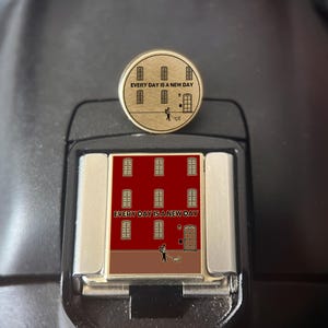 May include: A gold-coloured pin with the text "EVERY DAY IS A NEW DAY" and a building illustration. The pin is circular and features a person walking a dog. The pin is set on a black surface.