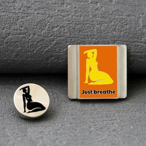 Pode incluir: Um berloque circular de metal prateado com a silhueta preta de uma figura sentada e um berloque retangular prateado com fundo laranja, uma silhueta amarela de uma figura sentada e o texto preto Just breathe.