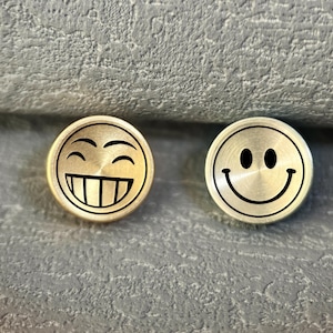May include: Two gold-coloured, circular objects with black smiley face designs. One has a wide open-mouth grin, the other a simple smile. They are set against a textured, grey background.