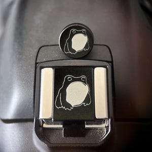 Puede incluir: Una tapa de zapata para cámara en color negro con una ilustración de rana grabada en blanco. La pieza superior circular muestra un diseño de rana a juego, mientras que la base rectangular encaja en la montura de la cámara.