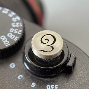 Pode incluir: Um bot&atilde;o disparador de c&acirc;mara circular de tom prateado com um n&uacute;mero tr&ecirc;s estilizado gravado. O bot&atilde;o est&aacute; montado na placa superior preta de uma c&acirc;mara com marcas de texto brancas vis&iacute;veis, incluindo velocidades do obturador e defini&ccedil;&otilde;es.