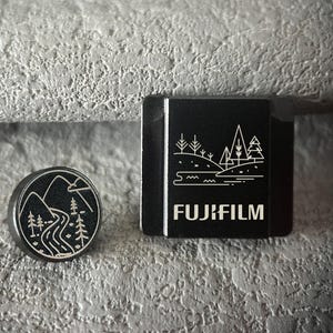 May include: A circular black metal pin featuring a white line art illustration of a winding river and mountains, positioned next to a rectangular black metal camera hot shoe cover with a white line art landscape and the text FUJIFILM.