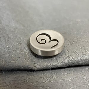 Pode incluir: Um pequeno bot&atilde;o circular de metal prateado escovado com um design de espiral gravado em preto na sua superf&iacute;cie superior. O bot&atilde;o tem uma forma de disco plano com um acabamento met&aacute;lico liso e um gr&aacute;fico de espiral escuro e estilizado.