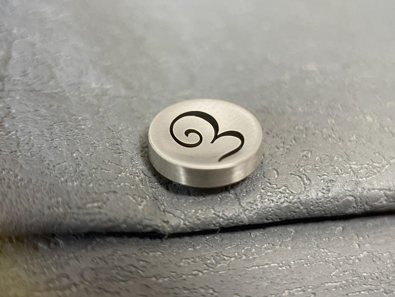 Pode incluir: Um pequeno bot&atilde;o circular de metal prateado escovado com um design de espiral gravado em preto na sua superf&iacute;cie superior. O bot&atilde;o tem uma forma de disco plano com um acabamento met&aacute;lico liso e um gr&aacute;fico de espiral escuro e estilizado.