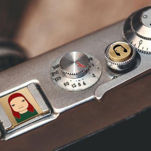 Puede incluir: Primer plano de una cámara plateada con múltiples diales y una pequeña imagen rectangular de un personaje de dibujos animados con pelo rojo y una camisa verde. Los diales de la cámara tienen números y marcas.