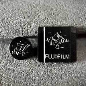 May include: Two black Fujifilm accessories with white mountain and tree designs. One is round, the other square with the Fujifilm logo. Both have a mountain and tree design.