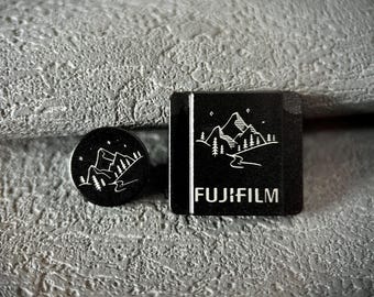 以下が含まれることがあります： 白い山と木の模様が入った2つの黒い富士フイルムアクセサリー。1つは丸く、もう1つは富士フイルムのロゴが入った四角です。どちらも山と木の模様があります。
