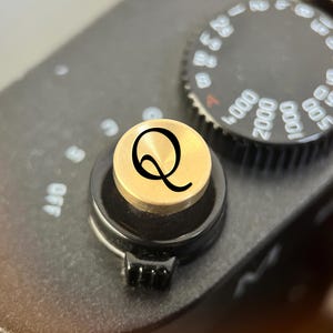 Pode incluir: Um bot&atilde;o disparador circular de metal dourado com uma letra Q preta gravada na superf&iacute;cie superior. O bot&atilde;o est&aacute; montado no corpo preto de uma c&acirc;mara ao lado de um mostrador preto com marca&ccedil;&otilde;es num&eacute;ricas brancas para a velocidade.