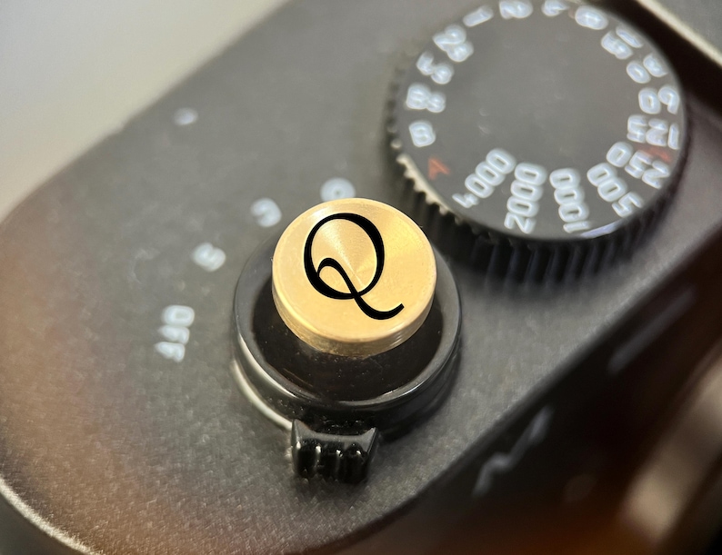 Pode incluir: Um bot&atilde;o disparador circular de metal dourado com uma letra Q preta gravada na superf&iacute;cie superior. O bot&atilde;o est&aacute; montado no corpo preto de uma c&acirc;mara ao lado de um mostrador preto com marca&ccedil;&otilde;es num&eacute;ricas brancas para a velocidade.