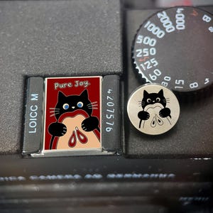 Pode incluir: Um broche retangular esmaltado com um gato preto comendo uma fatia de maçã com o texto Pure Joy, ao lado de um broche circular de metal prateado representando um gato preto segurando uma fatia de maçã, sobre um corpo de câmera preto.